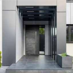 Nowoczesne wejście do domu z grafitowymi drzwiami z kamiennym panelem i długim uchwytem, zadaszone konstrukcją z metalowych belek. Obok donica z zielenią.