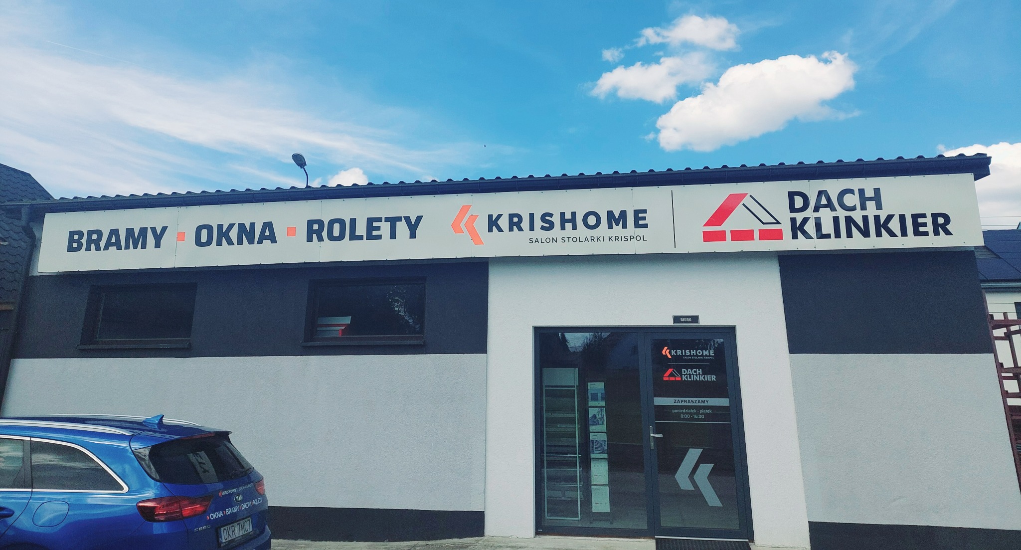 Fasada budynku z szyldem 'Bramy, Okna, Rolety - Krisshome Salon Stolarki Krispol Dach Klinkier'. Niebieski samochód zaparkowany przed budynkiem. Błękitne niebo z chmurami w tle.