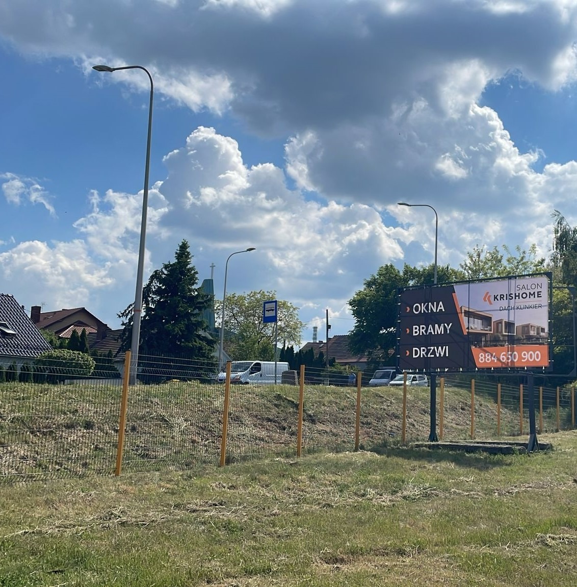 Billboard reklamowy firmy KrisHome prezentujący ofertę okien, bram i drzwi na tle błękitnego nieba z chmurami i zabudowań mieszkalnych.