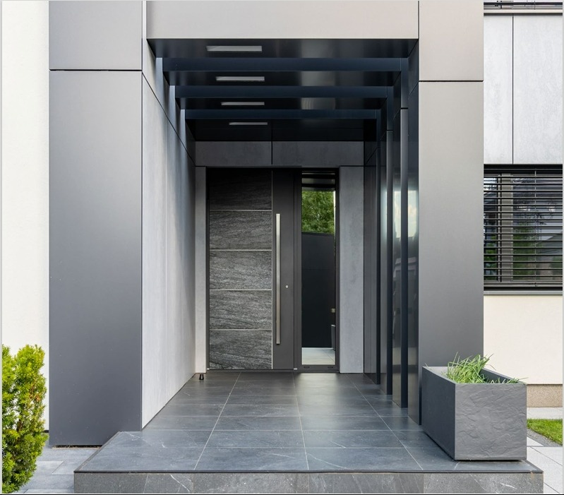 Nowoczesne wejście do domu z grafitowymi drzwiami z kamiennym panelem i długim uchwytem, zadaszone konstrukcją z metalowych belek. Obok donica z zielenią.
