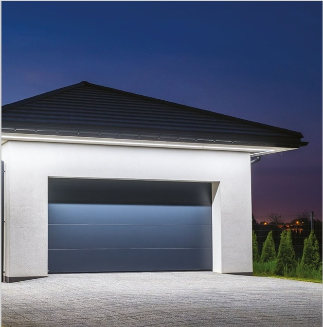 Nowoczesna brama garażowa segmentowa w kolorze antracytowym, oświetlona listwą LED, na tle nocnego nieba i zadbanego podjazdu z kostki brukowej.