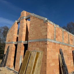 Błaszczuk budowlany zakład usługowy - Surowy stan domu z czerwonej cegły, widoczne ściany i otwory okienne, elementy betonowe, deski szalunkowe i błękitne niebo w tle. Dom w trakcie budowy.