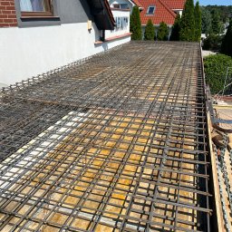 Błaszczuk budowlany zakład usługowy - Zbrojenie stropu na deskowaniu, widok z góry, Wołczkowo. Siatka z prętów stalowych przygotowana pod wylewkę betonową. Fragment budynku w tle.