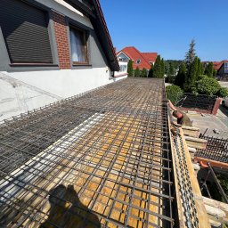 Błaszczuk budowlany zakład usługowy - Zbrojenie stropu domu jednorodzinnego z widoczną konstrukcją szalunkową i zbrojarzem przy pracy, w tle domy z czerwonymi dachami i błękitne niebo.