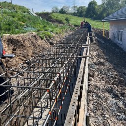 Błaszczuk budowlany zakład usługowy - Zbrojenie fundamentów w wykopie z deskowaniem, widoczni pracownicy. W tle zielony teren i fragment domu w stanie surowym zamkniętym.