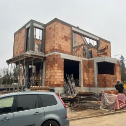 Błaszczuk budowlany zakład usługowy - Dwukondygnacyjny dom w trakcie budowy z czerwonej cegły Porotherm, widoczne szalunki okienne, wsporniki stropowe i auto na pierwszym planie. Szary dzień.