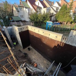 Błaszczuk budowlany zakład usługowy - Fundamenty zabezpieczone brązową izolacją bitumiczną, widok z góry na dół, drabina, zbrojenie, częściowo widoczna osoba w ubraniu roboczym, w tle zabudowa miejska.