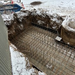 Błaszczuk budowlany zakład usługowy - Zbrojenie fundamentu w wykopie, częściowo zasypanym śniegiem. Widoczne stalowe pręty i siatka zbrojeniowa. Obok studzienka kanalizacyjna i kontener na odpady.