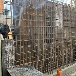 Błaszczuk budowlany zakład usługowy - Zbrojenie pionowej ściany szalunkowej z widocznymi elementami mocującymi i deskami szalunkowymi w dolnej części. W tle widoczne materiały budowlane i drzewo.