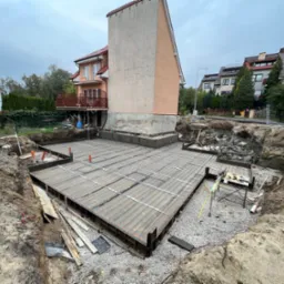 Zbrojenie fundamentów pod budynek mieszkalny, widok z góry. Stalowa siatka zbrojeniowa ułożona w wykopie, w tle dom w trakcie budowy i zabudowa jednorodzinna.