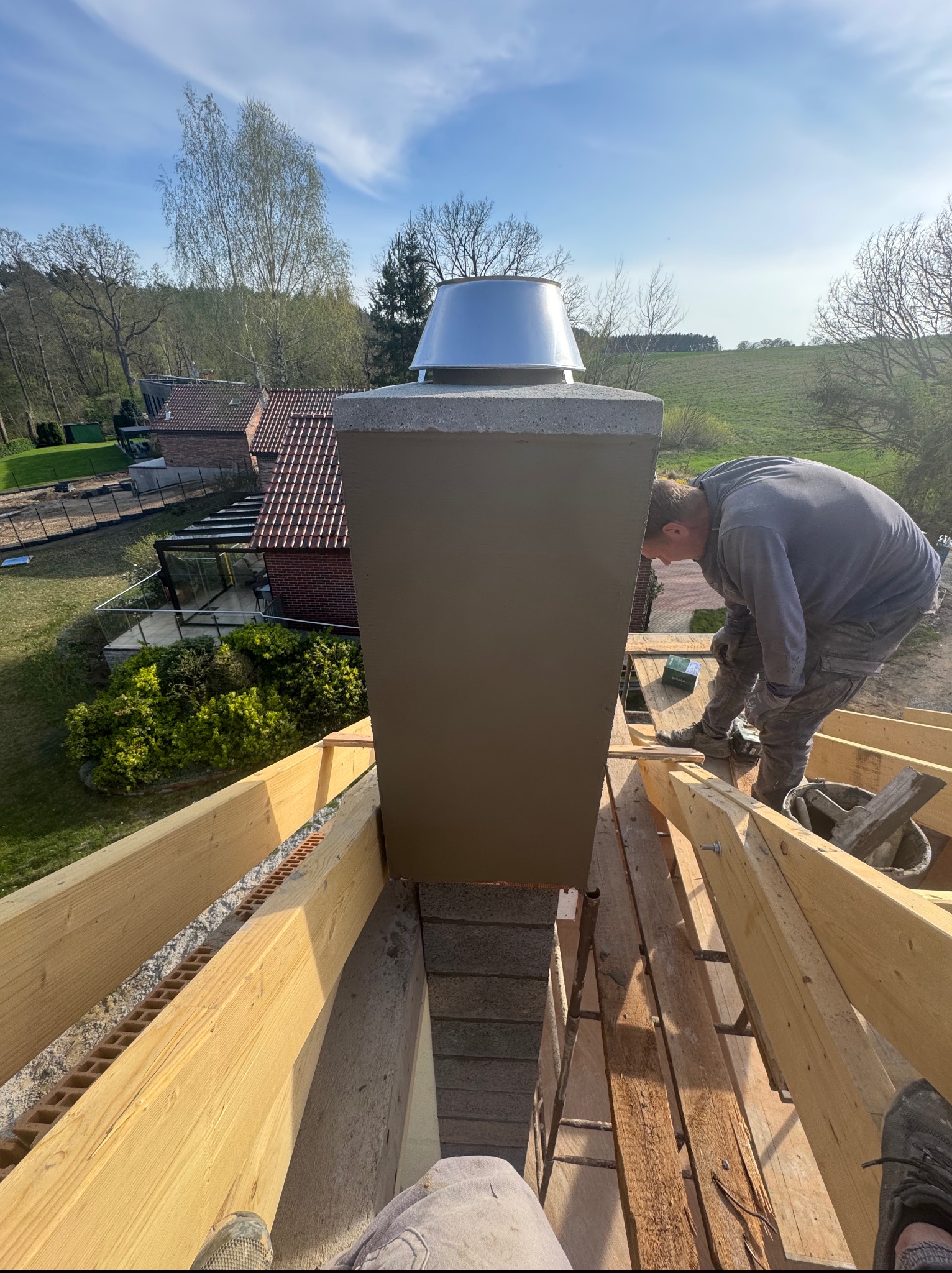 Murarz podczas pracy nad kominem na dachu domu w słoneczny dzień. Widoczne drewniane elementy konstrukcji dachu i narzędzia murarskie.