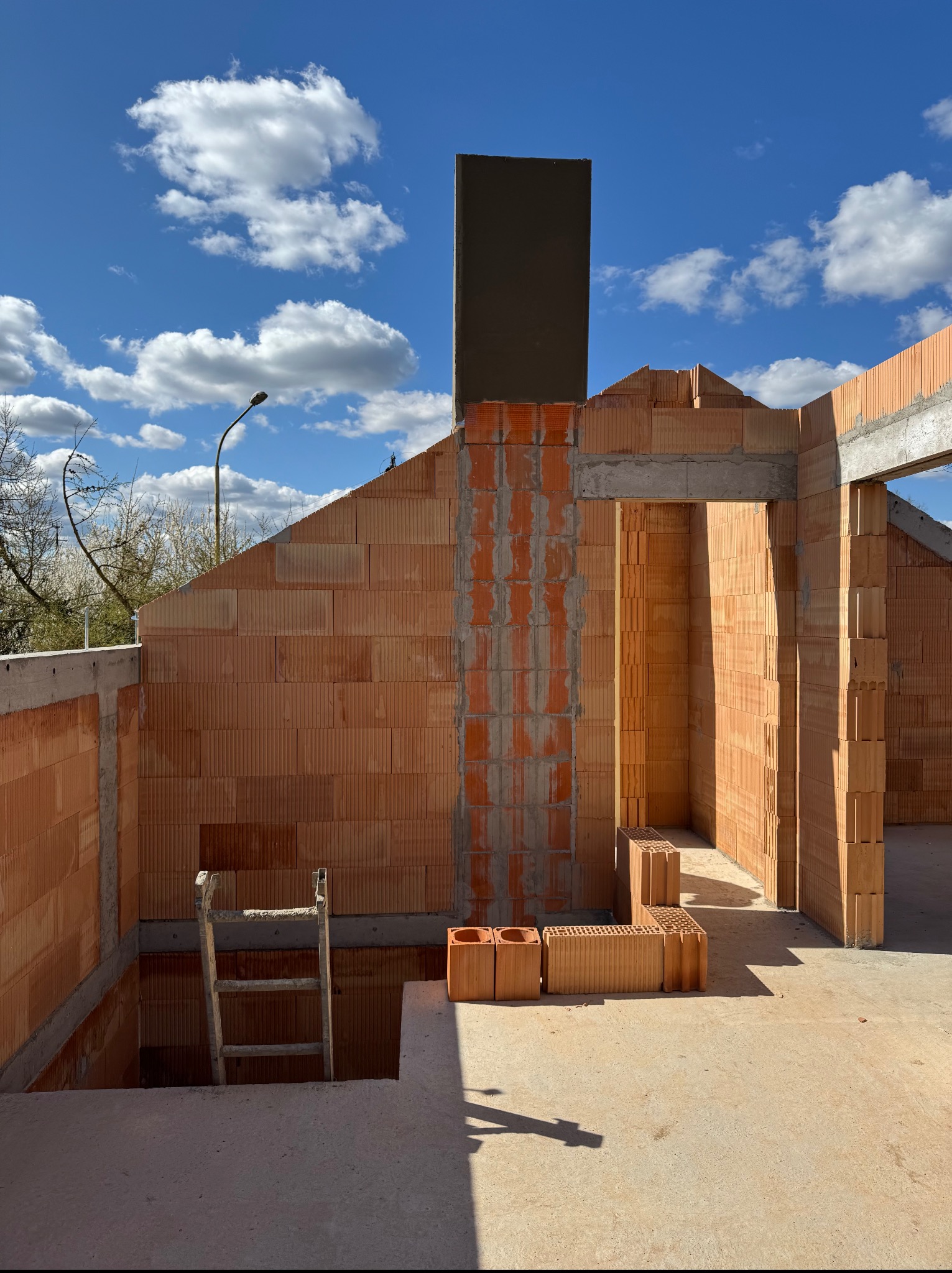 Wnętrze domu w trakcie murowania ścian z czerwonej cegły ceramicznej. Widoczne fragmenty ścian, otwory okienne i drzwiowe, drabina. Błękitne niebo z chmurami w tle.