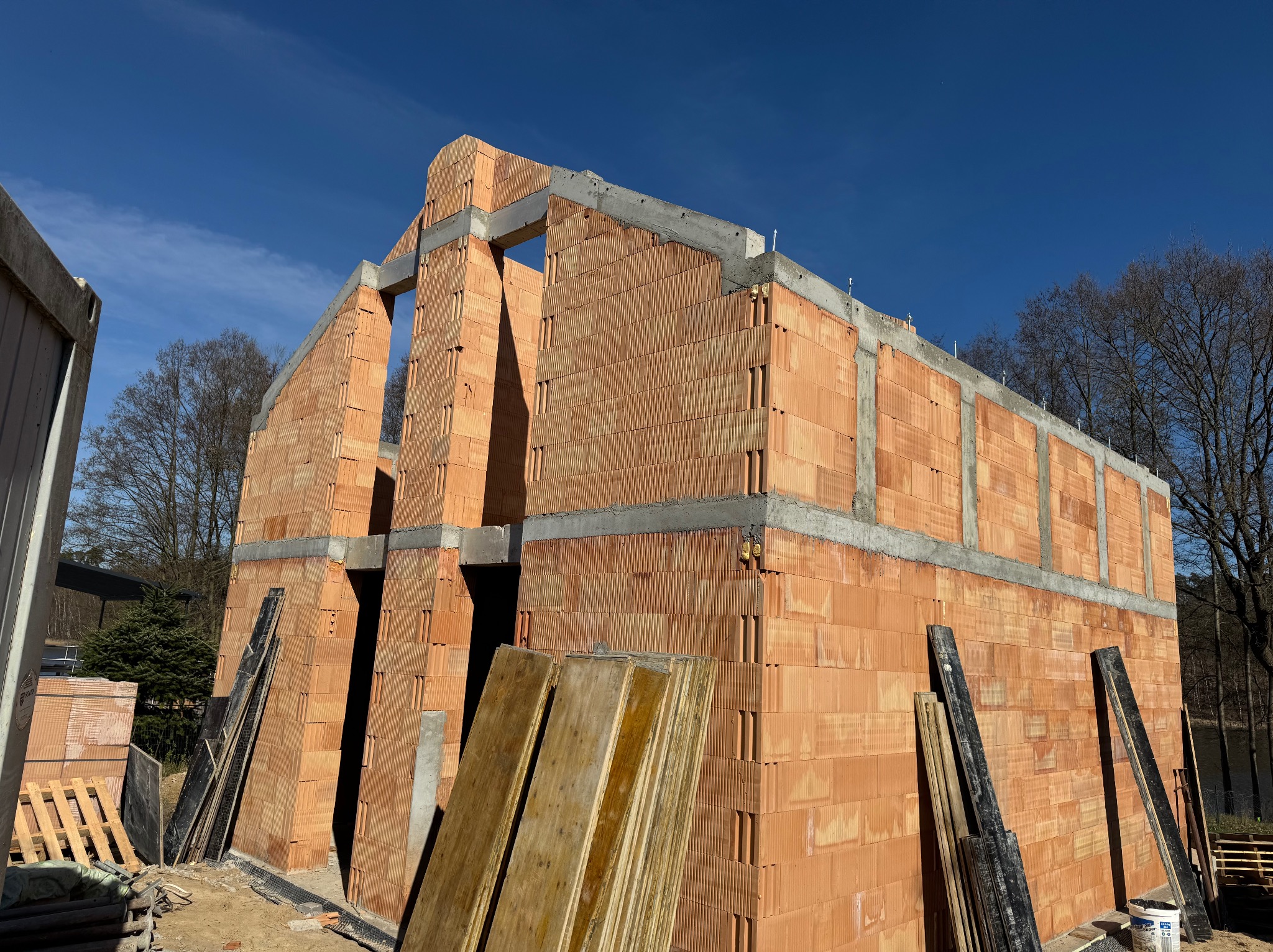 Surowy stan domu z czerwonej cegły, widoczne ściany i otwory okienne, elementy betonowe, deski szalunkowe i błękitne niebo w tle. Dom w trakcie budowy.