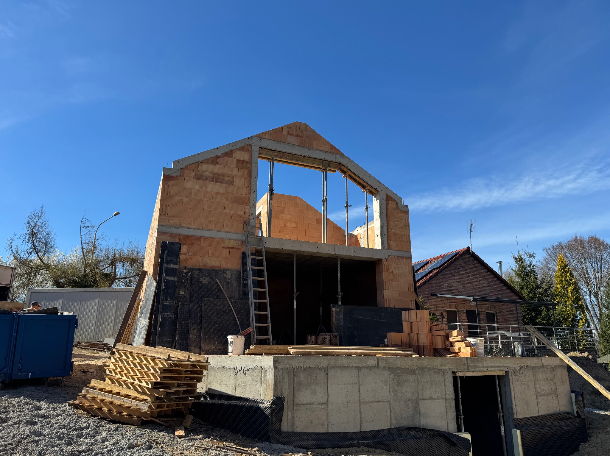 Surowy stan domu w budowie: fundament z betonu, ściany z czerwonej cegły, drewniane podpory. Widoczne materiały budowlane i otoczenie placu budowy w słoneczny dzień.