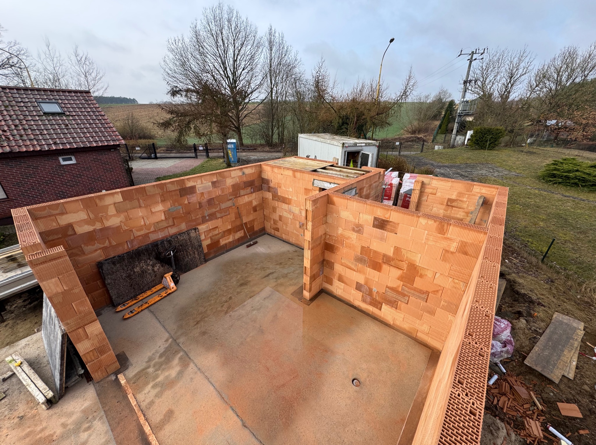 Wysoki kąt: Budowa domu z czerwonej ceramiki porotherm, ściany wzniesione do pewnej wysokości. Widoczne palety z materiałem, otoczenie działki, pochmurne niebo.