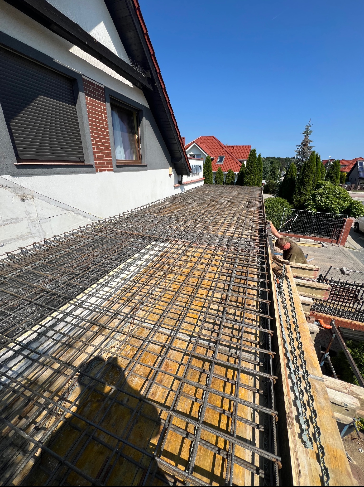 Zbrojenie stropu domu jednorodzinnego z widoczną konstrukcją szalunkową i zbrojarzem przy pracy, w tle domy z czerwonymi dachami i błękitne niebo.