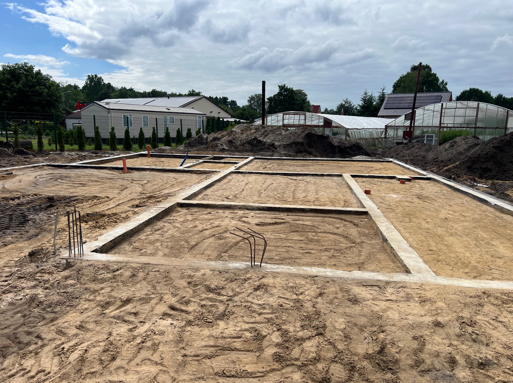Zalane fundamenty pod budowę domu, widoczne wystające pręty zbrojeniowe. Na działce w tle dom modułowy i szklarnie. Pochmurne niebo.