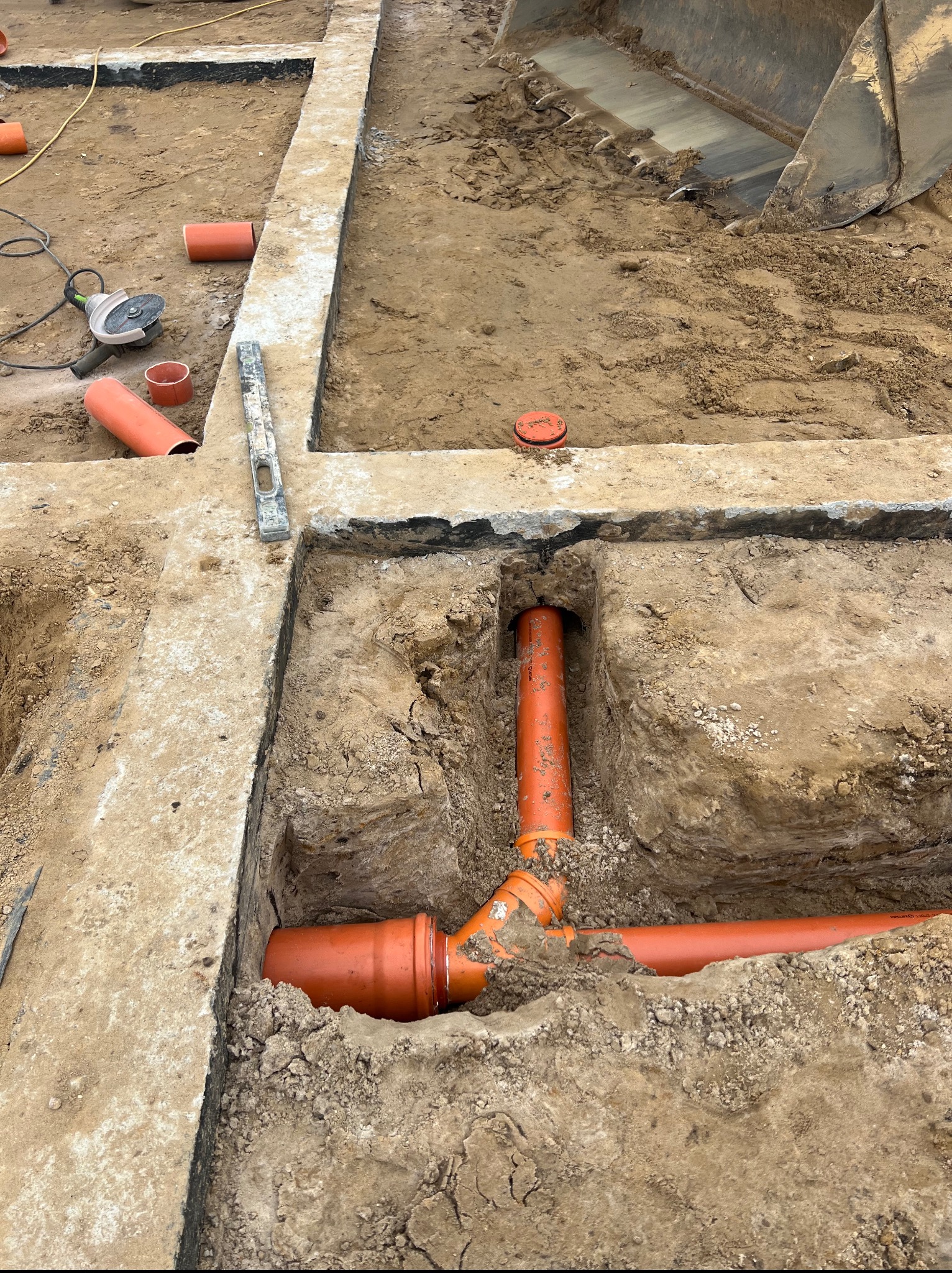 Fundamenty z wykopem pod instalacje: pomarańczowe rury kanalizacyjne w ziemi, poziomica, szlifierka kątowa i łyżka koparki w tle. Prace ziemne.