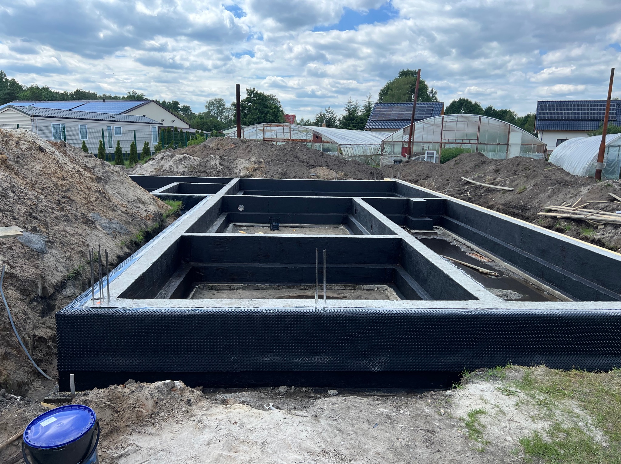 Zabezpieczone fundamenty domu jednorodzinnego w trakcie budowy, izolacja pionowa, betonowe ściany fundamentowe, widoczne zbrojenia, ziemia i budynki w tle.