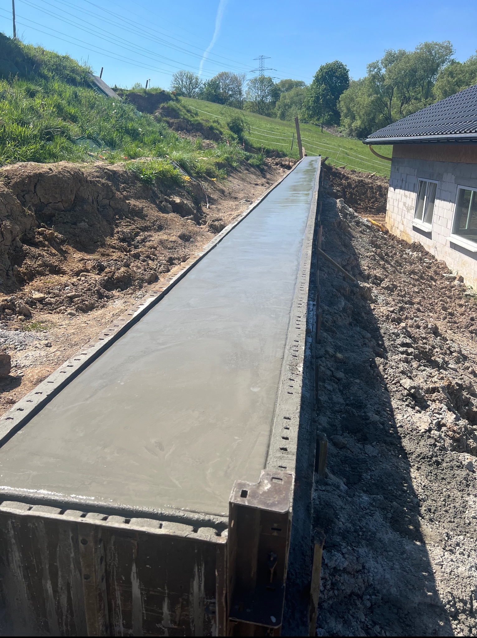 Wylany fundament pod budynek w szalunku, widok z góry. Świeży beton, ziemia i fragment ściany budynku w tle. Słoneczny dzień.