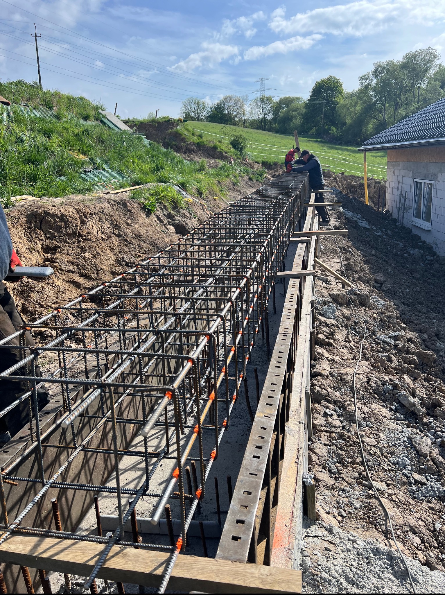 Zbrojenie fundamentów w wykopie z deskowaniem, widoczni pracownicy. W tle zielony teren i fragment domu w stanie surowym zamkniętym.