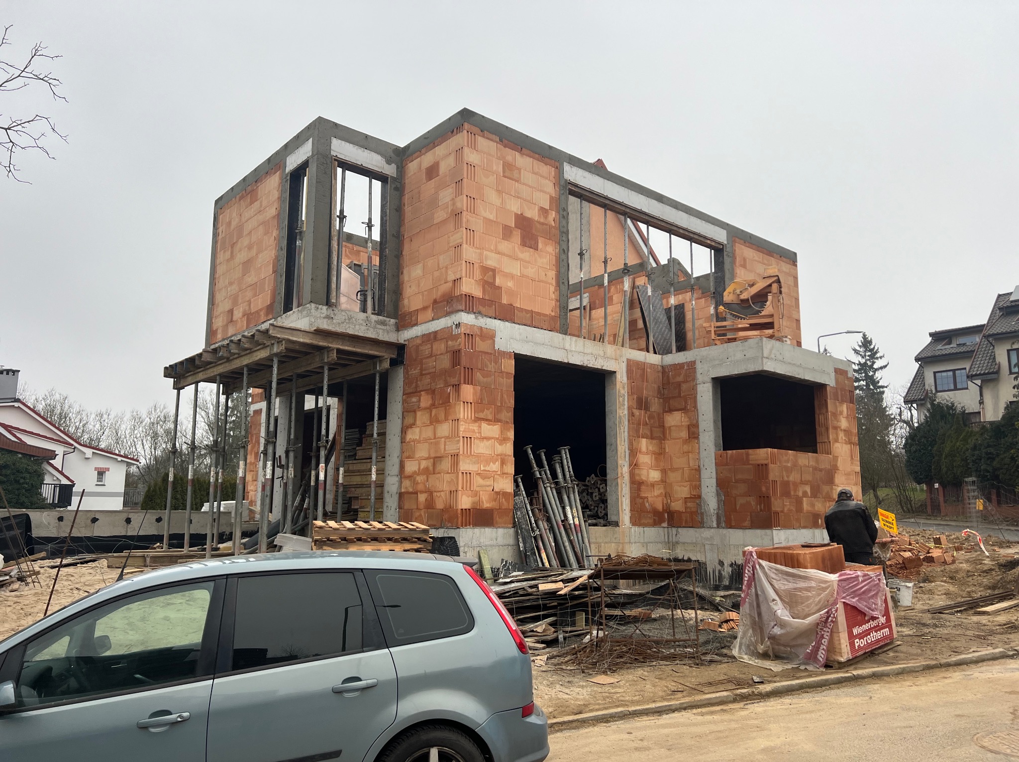 Dwukondygnacyjny dom w trakcie budowy z czerwonej cegły Porotherm, widoczne szalunki okienne, wsporniki stropowe i auto na pierwszym planie. Szary dzień.