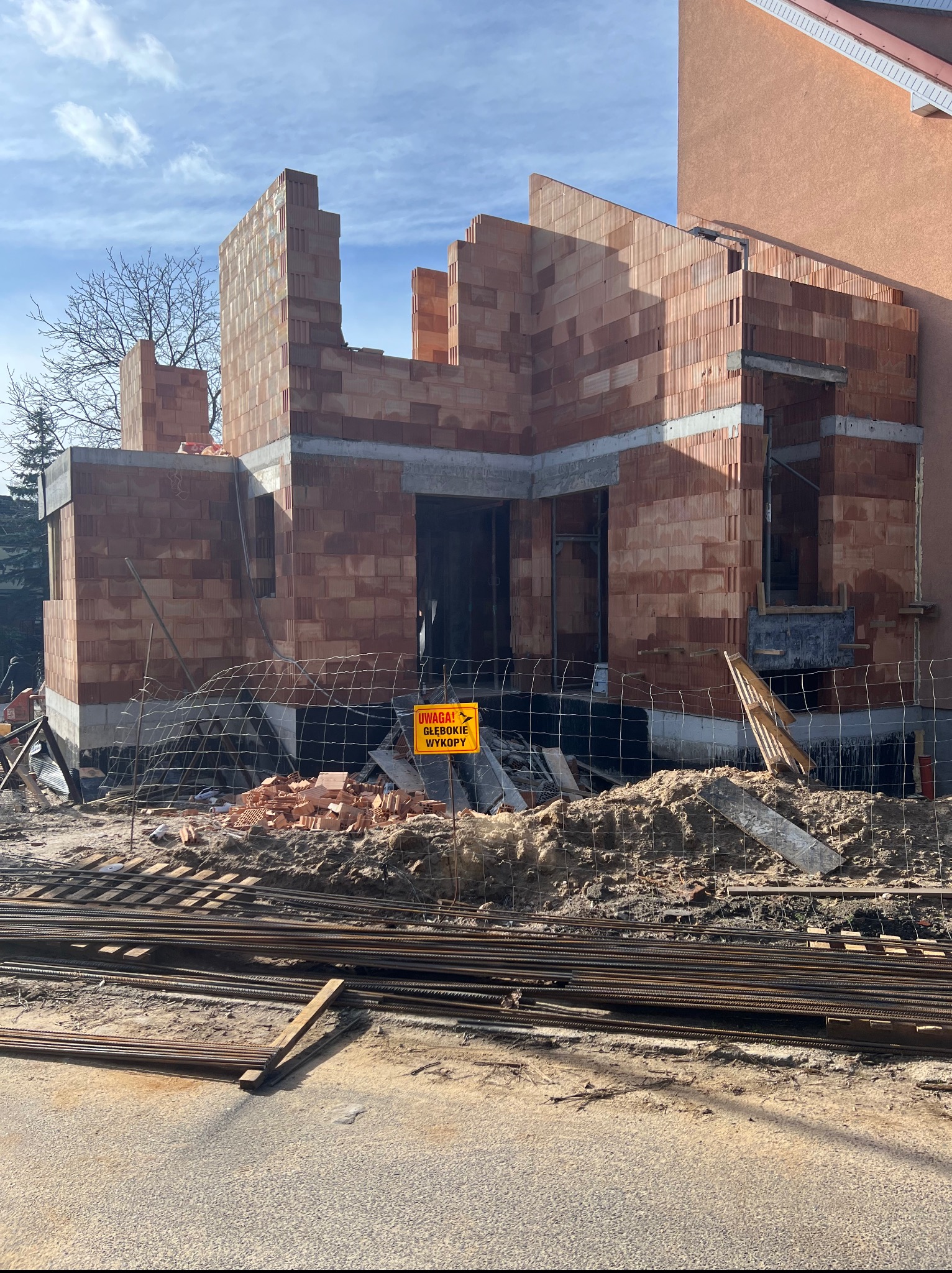 Wznoszenie ścian domu z czerwonej cegły, widoczne zbrojenia i tablica ostrzegawcza. Częściowo wybudowany budynek w trakcie prac murarskich.