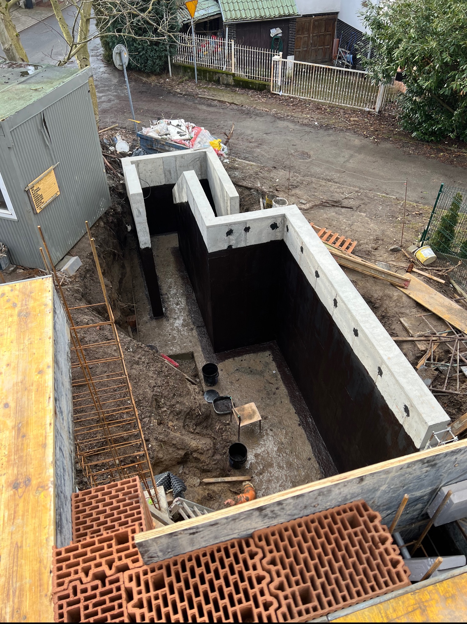 Fundamenty domu w trakcie budowy: wykop, izolacja bitumiczna ścian, betonowy wieniec. Widok z góry na plac budowy z cegłami i drabiną.