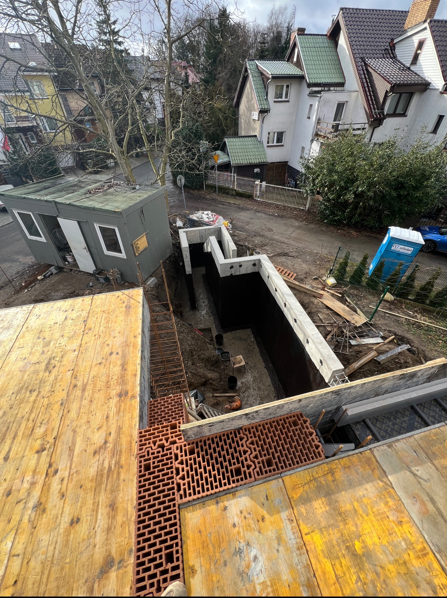 Fundamenty domu w trakcie budowy, widok z góry na wykop z deskowaniem i zbrojeniem. Na pierwszym planie cegły i elementy drewniane szalunku. W tle budynki mieszkalne.