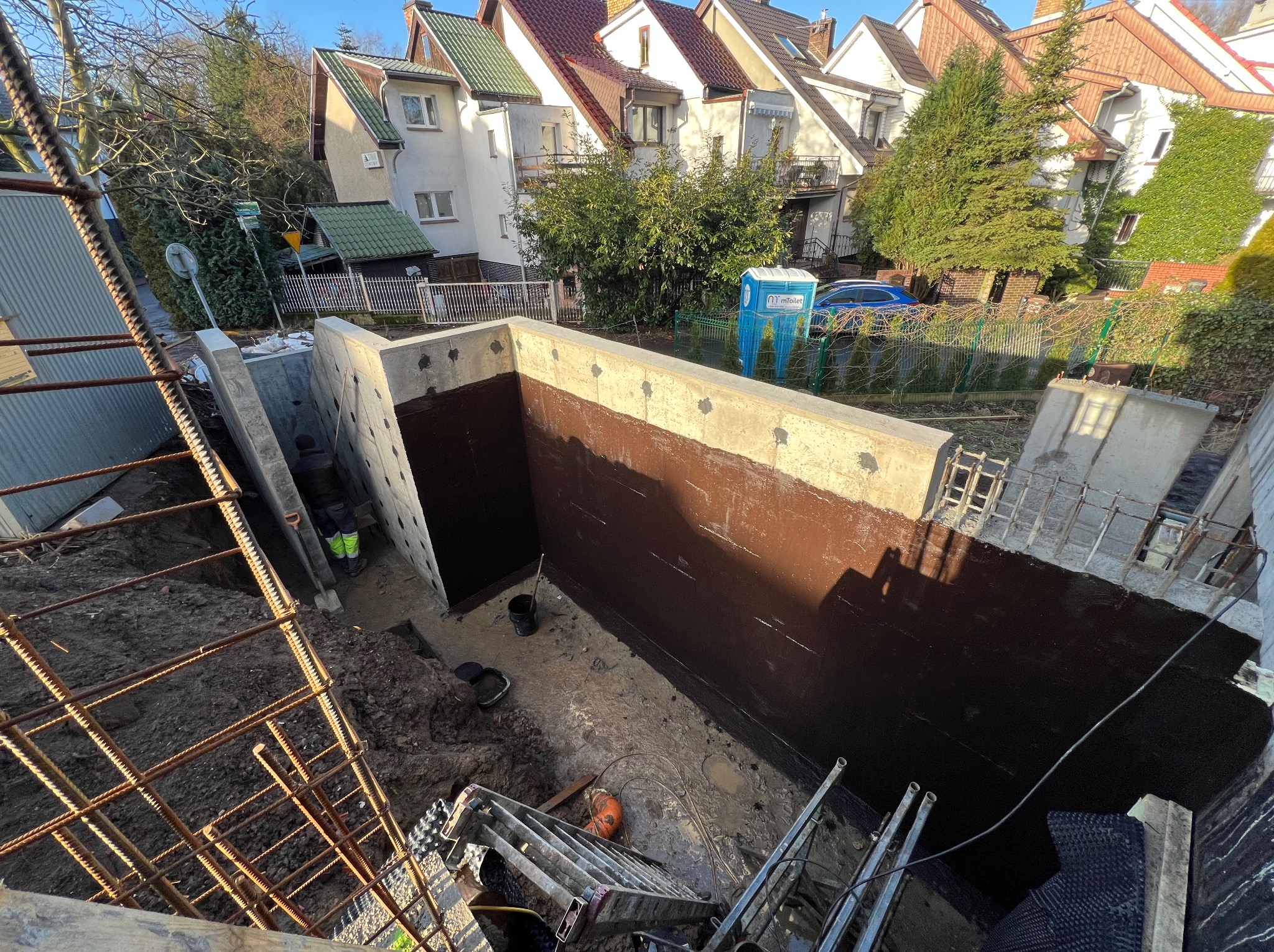 Fundamenty zabezpieczone brązową izolacją bitumiczną, widok z góry na dół, drabina, zbrojenie, częściowo widoczna osoba w ubraniu roboczym, w tle zabudowa miejska.