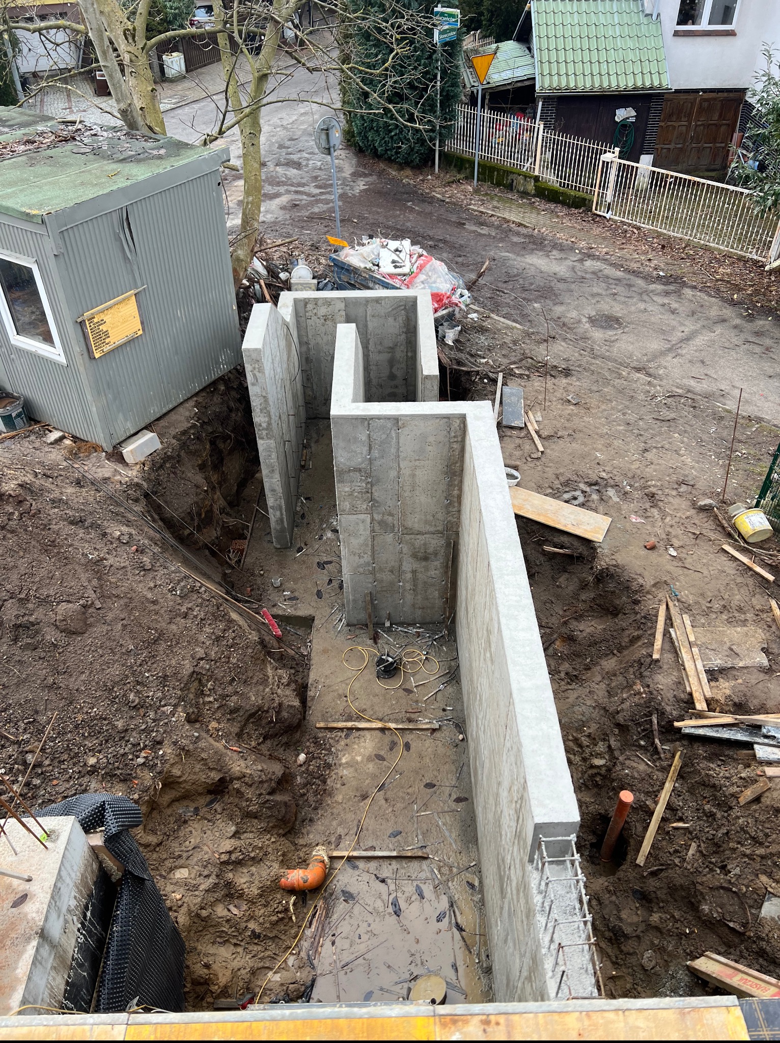 Betonowe ściany fundamentowe w wykopie, widok z góry. Elementy zbrojenia wystają z jednej ze ścian. Obok barak budowlany i ulica.