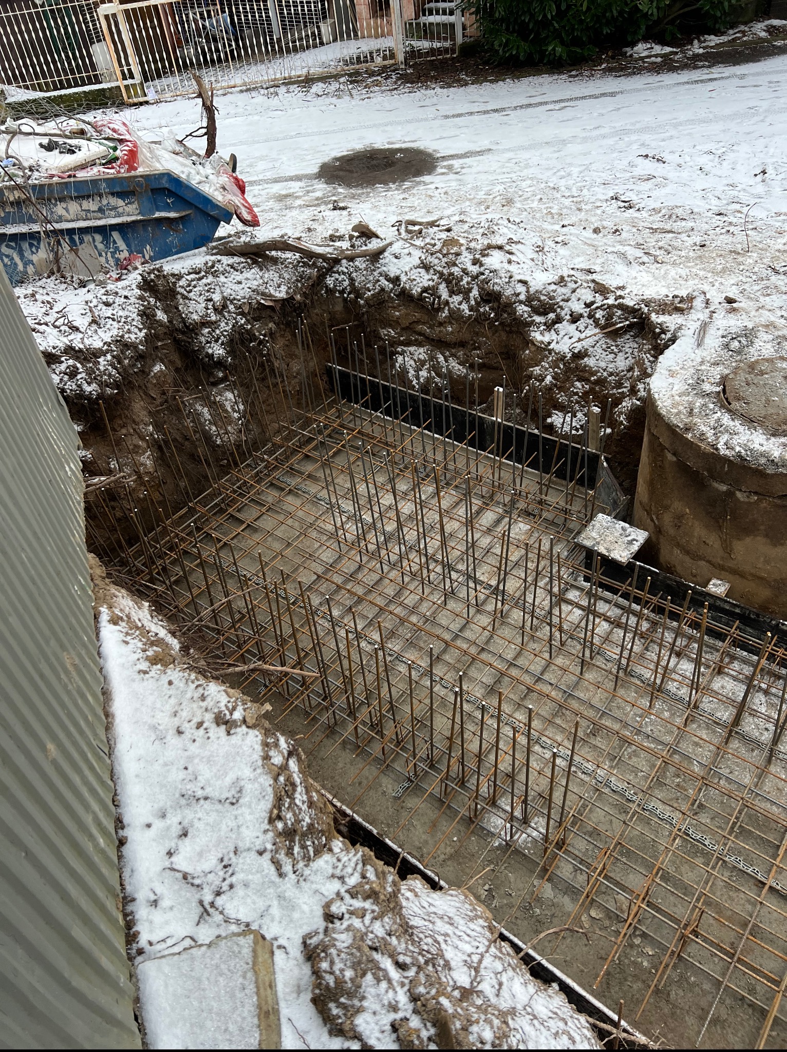 Zbrojenie fundamentu w wykopie, częściowo zasypanym śniegiem. Widoczne stalowe pręty i siatka zbrojeniowa. Obok studzienka kanalizacyjna i kontener na odpady.