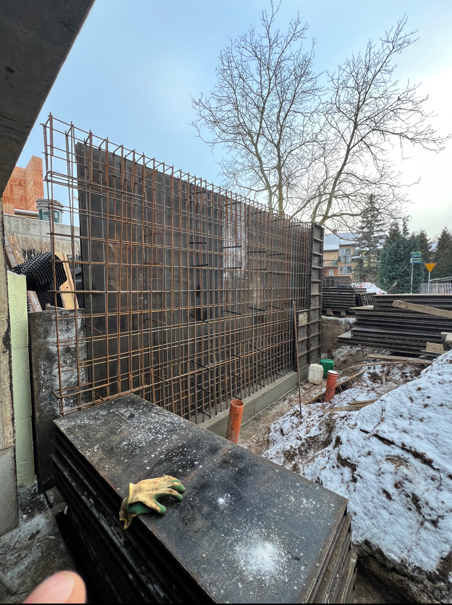 Zbrojenie fundamentów z deskowaniem i rurami kanalizacyjnymi na placu budowy. Na pierwszym planie stos płyt i robocza rękawica. W tle budynki i drzewa.