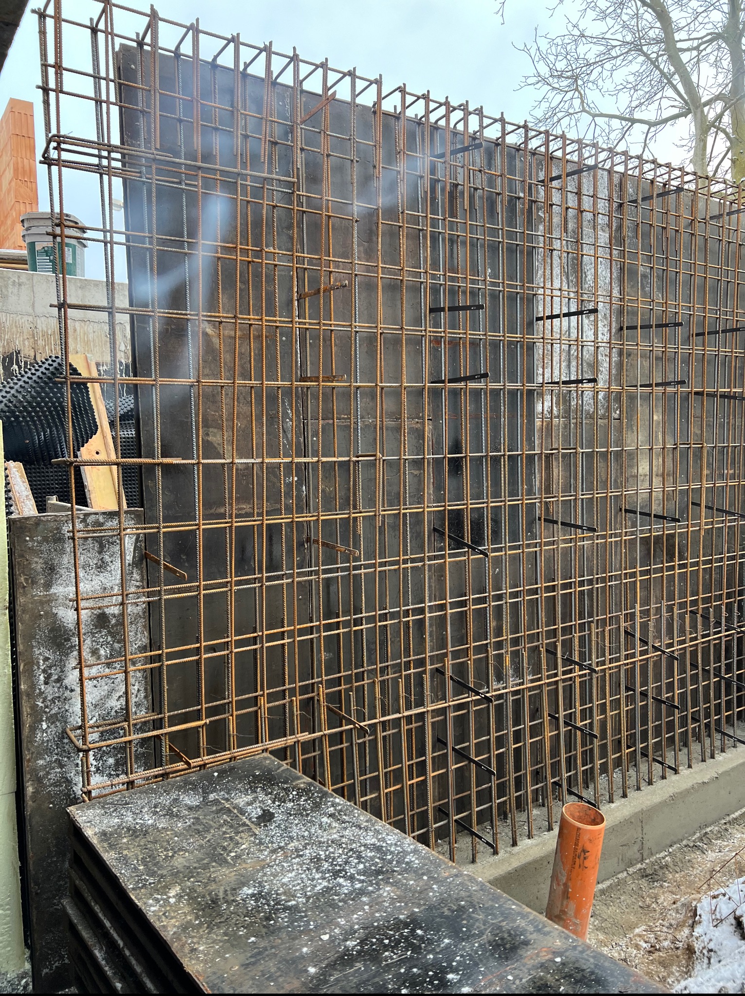 Zbrojenie pionowej ściany szalunkowej z widocznymi elementami mocującymi i deskami szalunkowymi w dolnej części. W tle widoczne materiały budowlane i drzewo.