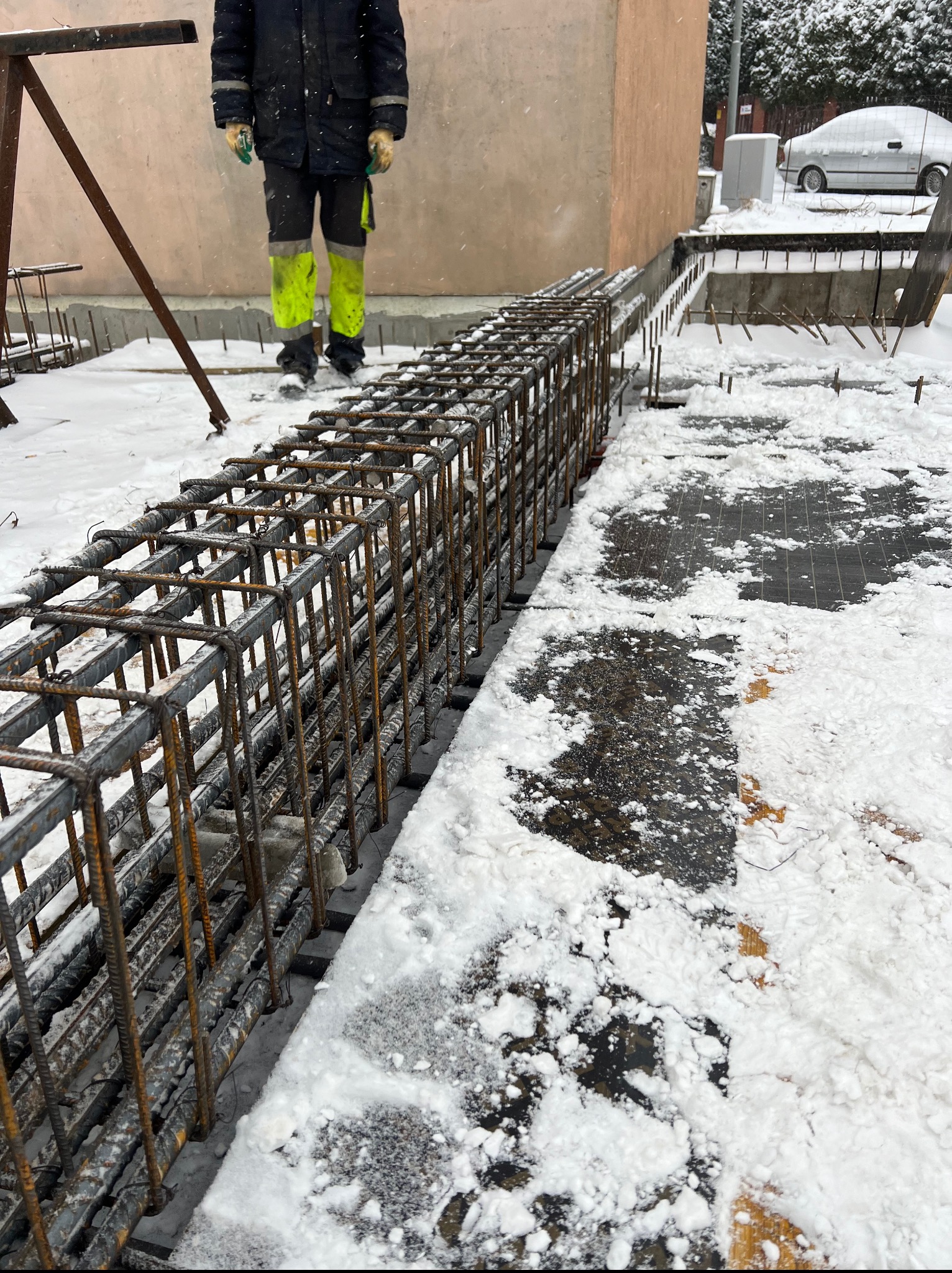 Zimowy kadr z budowy: zbrojenie fundamentów w śniegu, widoczny fragment budynku i pracownik w odblaskowej odzieży. Ujęcie z niskiej perspektywy, podkreślające detale konstrukcji.