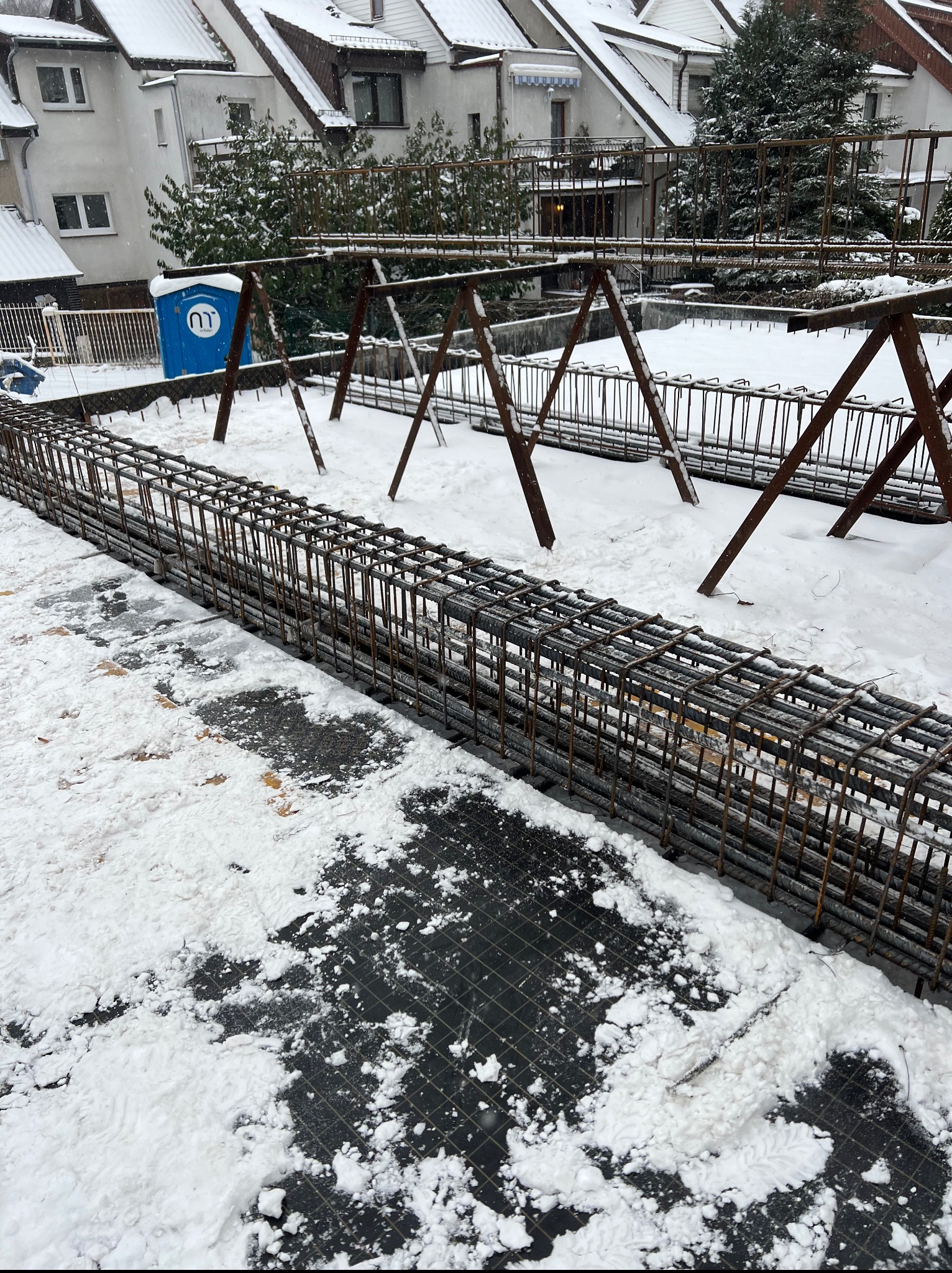 Zimowy widok na zbrojenie fundamentów pod budynek. Metalowa konstrukcja w śniegu, w tle budynki mieszkalne i przenośna toaleta.
