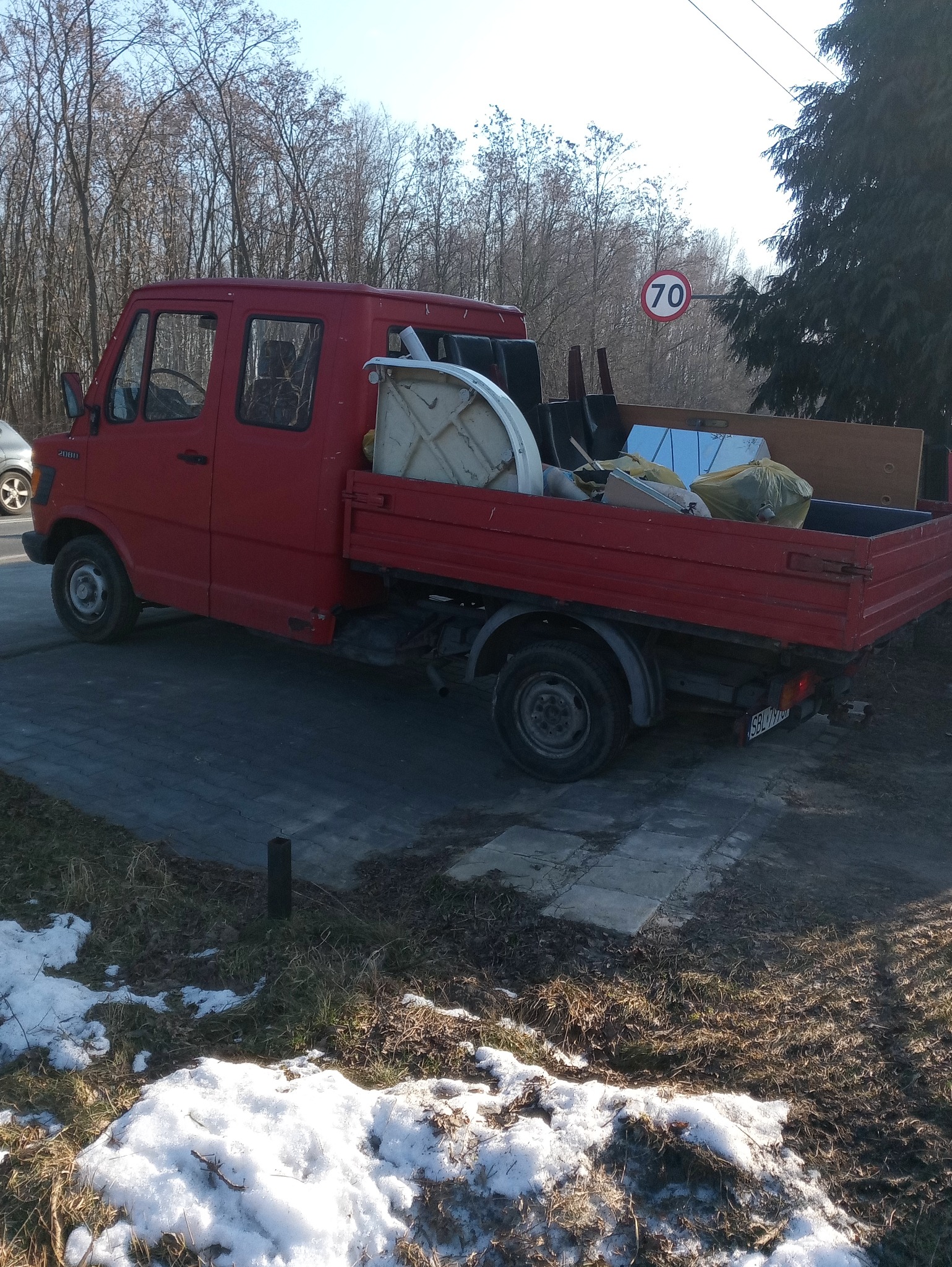 Czerwony samochód dostawczy z otwartą skrzynią załadowaną różnymi przedmiotami, w tle drzewa i znak drogowy z ograniczeniem prędkości do 70.