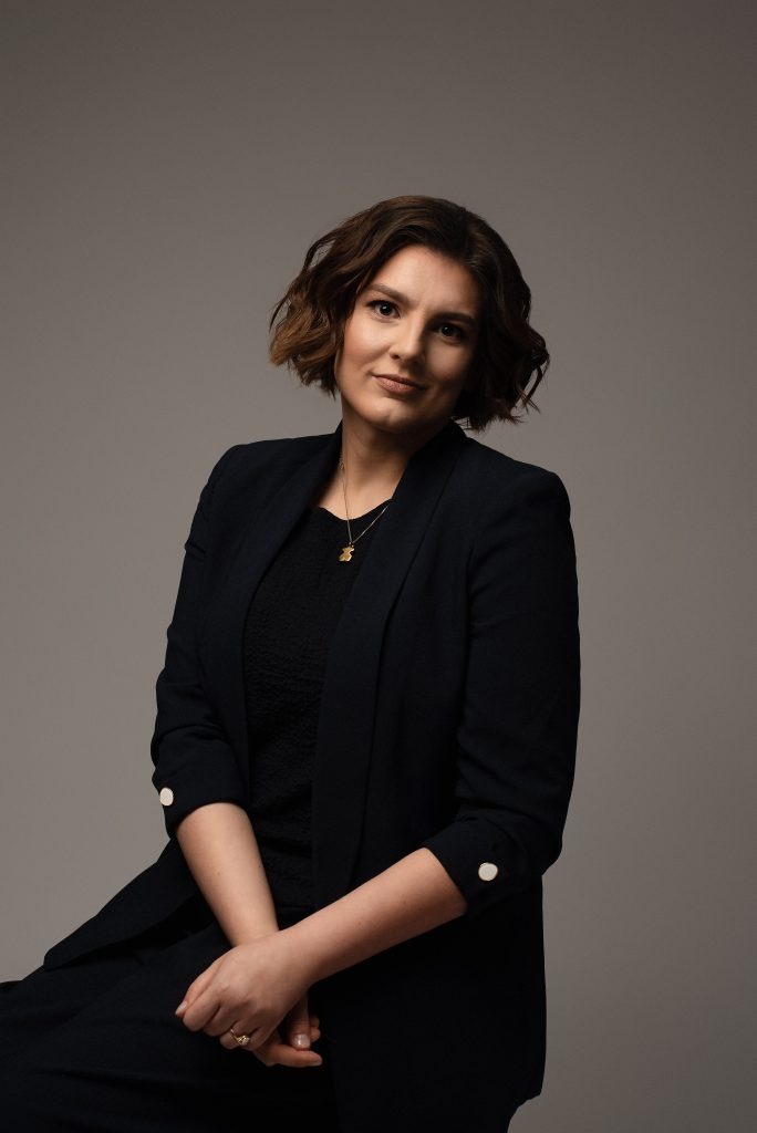 Portret kobiety w czarnym żakiecie z ozdobnymi guzikami, na szyi złoty naszyjnik z zawieszką misia, na szarym tle. Profesjonalny wizerunek.