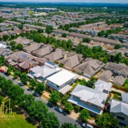 Widok z lotu ptaka na osiedle domów jednorodzinnych z zielenią i ulicami. Domy o różnej architekturze i kolorystyce dachów. Na pierwszym planie logo firmy Aurevia Nieruchomości.