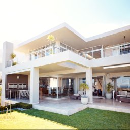 AUREVIA INVESTMENTS SPÓŁKA Z OGRANICZONĄ ODPOWIEDZIALNOŚCIĄ - Dwupiętrowa, biała willa z balkonem i dużymi oknami wychodzącymi na trawnik. Widoczne meble ogrodowe i elementy dekoracyjne. Minimalistyczny styl.