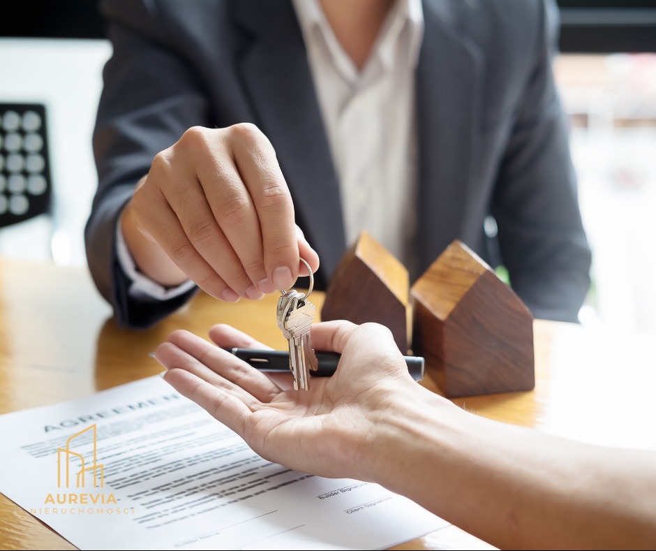 Przekazanie kluczy do mieszkania na umowie. W tle drewniane domki, symbol transakcji nieruchomości. Elegancki garnitur, jasne wnętrze.