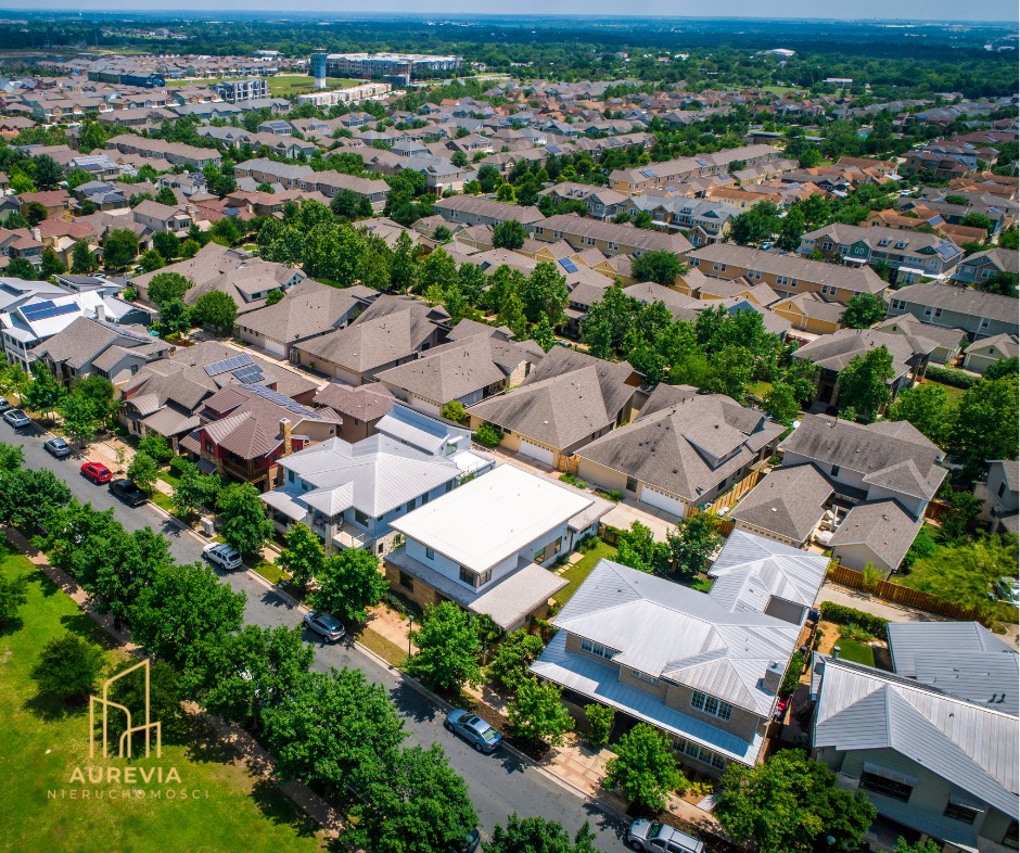 Widok z lotu ptaka na osiedle domów jednorodzinnych z zielenią i ulicami. Domy o różnej architekturze i kolorystyce dachów. Na pierwszym planie logo firmy Aurevia Nieruchomości.