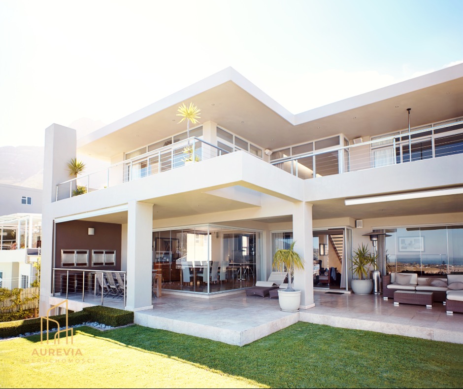 Dwupiętrowa, biała willa z balkonem i dużymi oknami wychodzącymi na trawnik. Widoczne meble ogrodowe i elementy dekoracyjne. Minimalistyczny styl.