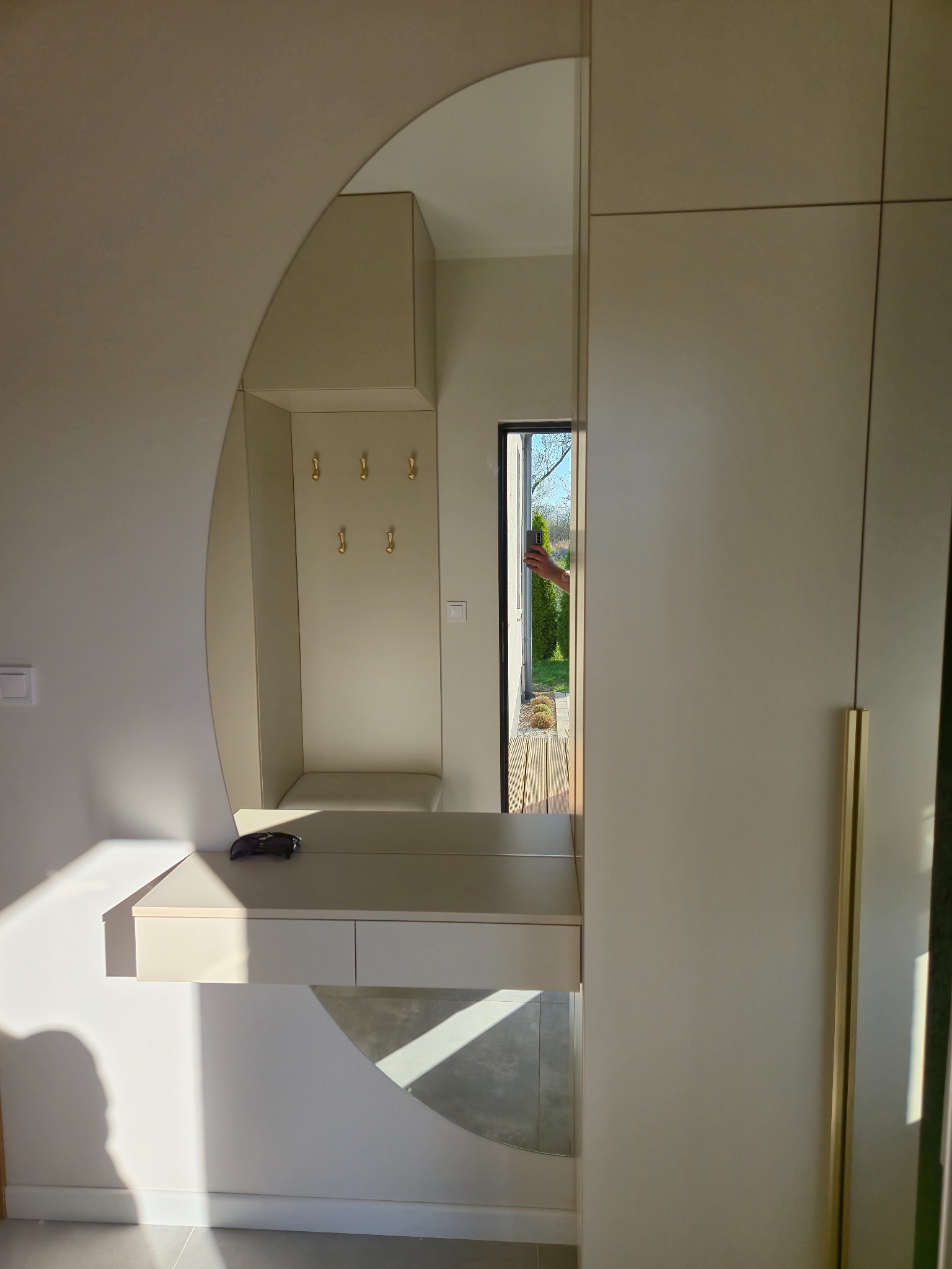 Beżowa szafa z lustrem i wnęką z wieszakami, element zabudowy przedpokoju. W lustrze odbicie ogrodu. Złote uchwyty, minimalistyczny design i światło słoneczne.