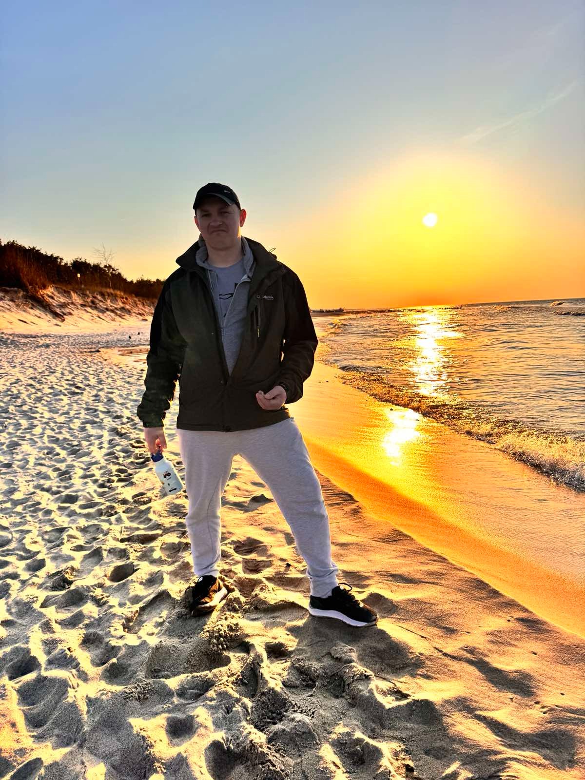 Mężczyzna w sportowym stroju stoi na piaszczystej plaży o zachodzie słońca, trzymając butelkę z wodą. Widoczne morze i niebo w ciepłych, pomarańczowych barwach.