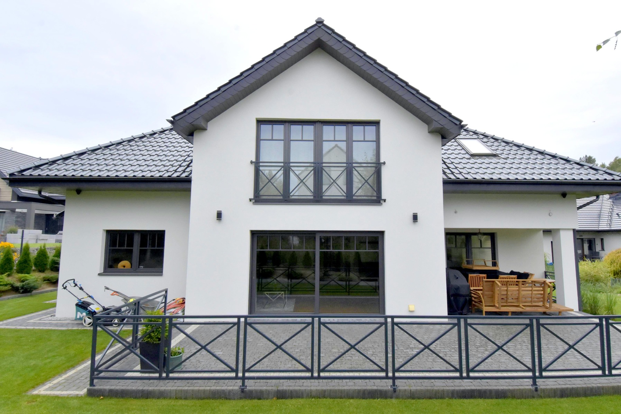 Nowoczesny dom jednorodzinny z czarną stolarką okienną i balkonem zabezpieczonym balustradą, widok z ogrodu. Na pierwszym planie metalowe ogrodzenie.