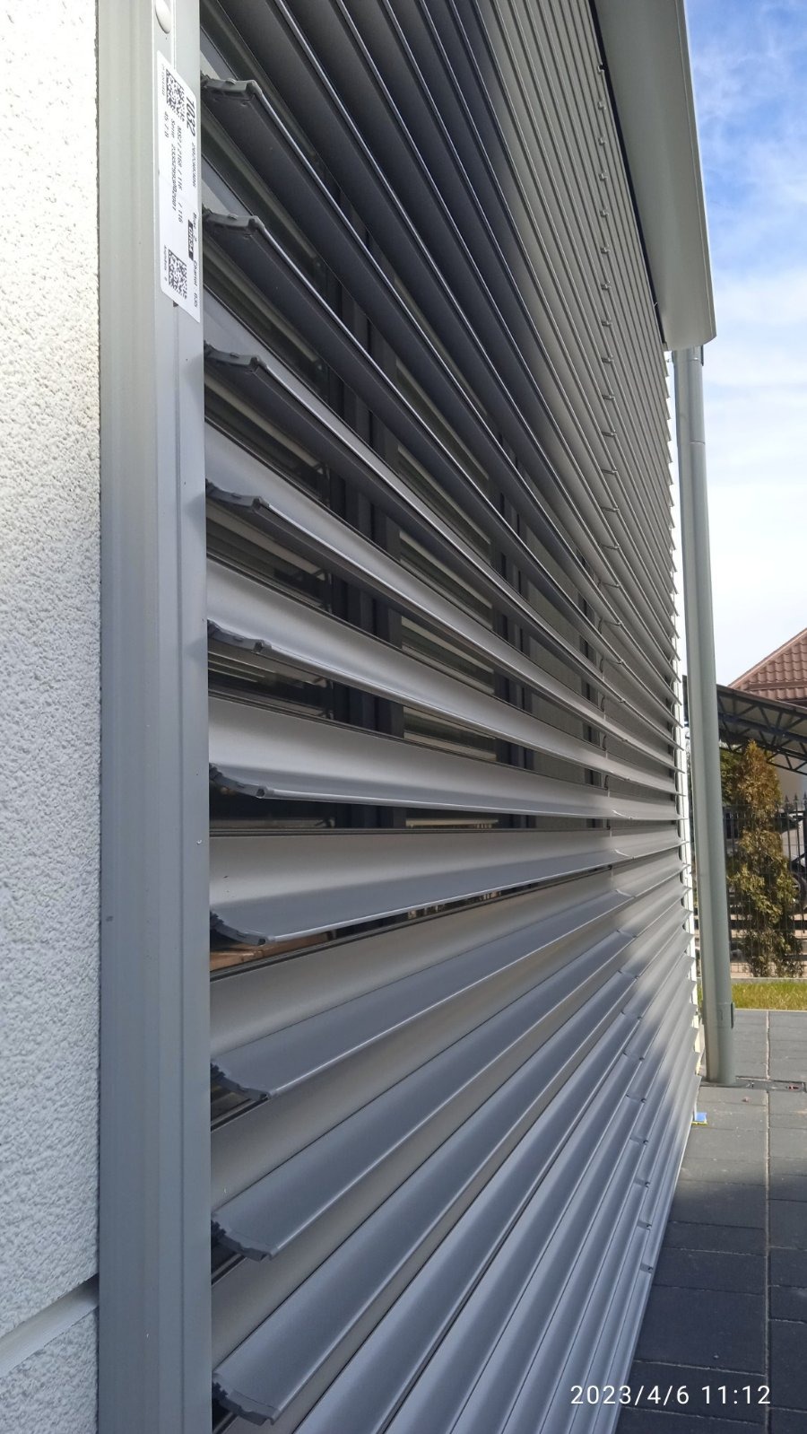 Zewnętrzna roleta aluminiowa w kolorze szarym, zamontowana na elewacji budynku. Widoczne lamele i system prowadnic. Na roletach naklejka z oznaczeniem modelu.