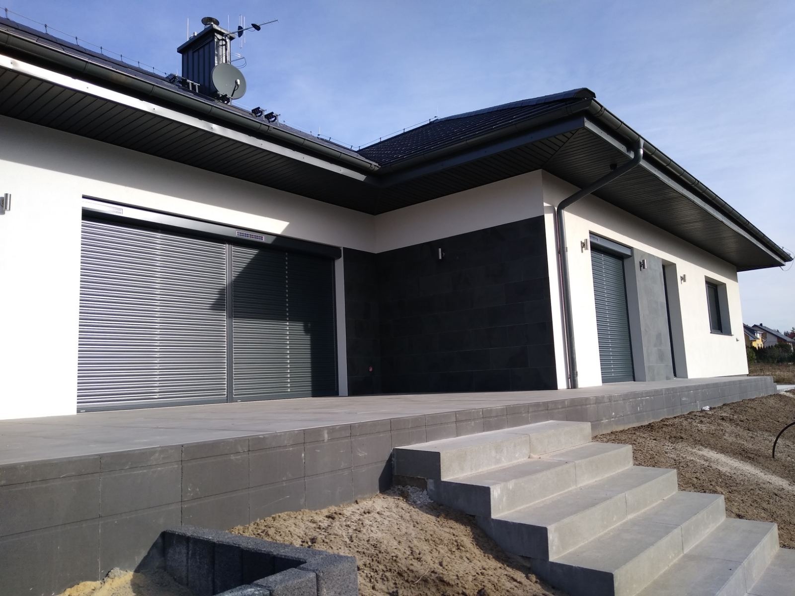 Nowoczesny dom z szarą elewacją i zewnętrznymi roletami antywłamaniowymi. Widoczne schody wejściowe i fragment ogrodu.