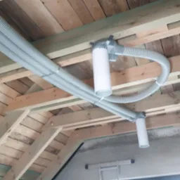 Montaż systemu rekuperacji pod drewnianym stropem poddasza. Widoczne szare rury karbowane i białe elementy systemu wentylacyjnego.