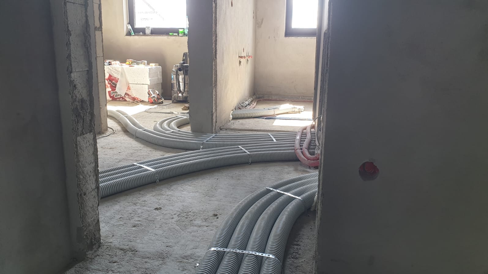 Szare rury wentylacyjne ułożone na podłodze w surowym wnętrzu, widoczne okno i materiały budowlane w tle, instalacja rekuperacji w trakcie montażu.